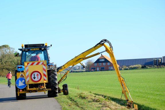 Gelber Traktor mit einem Ausleger schneidet Gras am Straßenrand auf einem Feld.