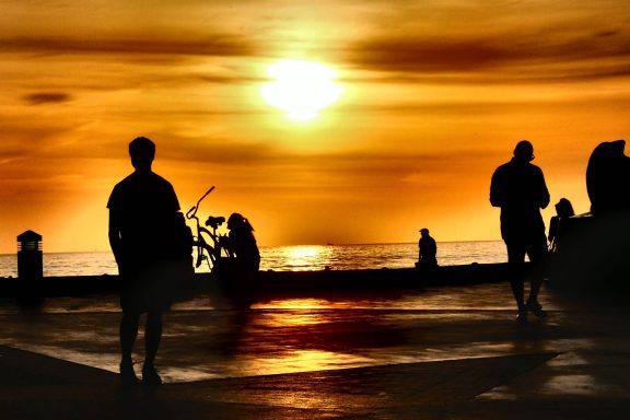 Silhouetten von Menschen bei Sonnenuntergang am Meer mit orangefarbenem Himmel.