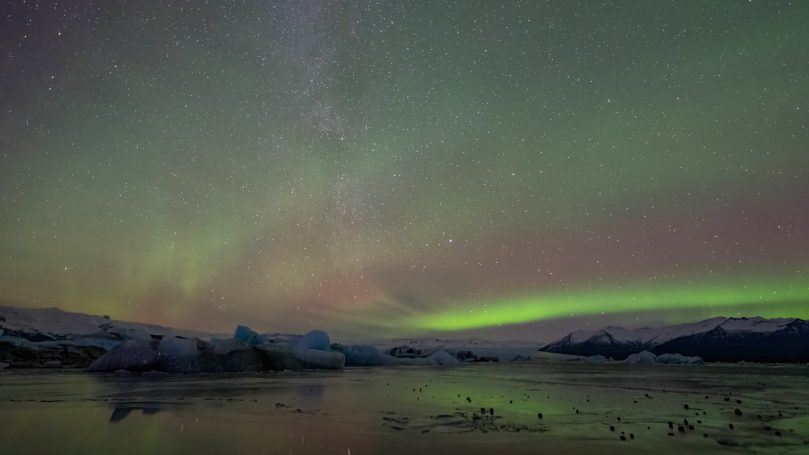 Nordlichter über einem ruhigen See mit Eisbergen im Vordergrund.