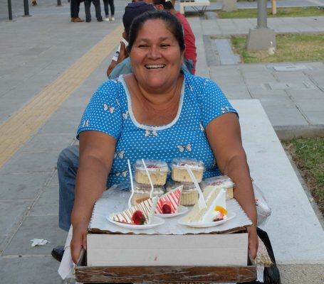 Eine lächelnde Frau verkauft Desserts auf einem Tablett in einer urbanen Umgebung.