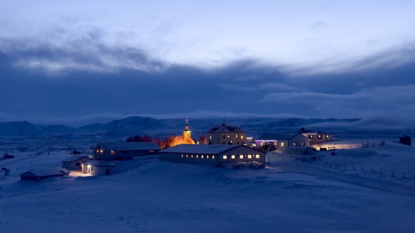 Winterliche Landschaft mit beleuchteten Gebäuden in der Ferne und schneebedecktem Boden.