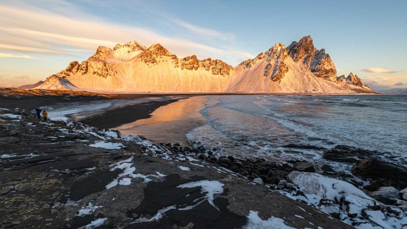 Schneebedeckte Berge neben einem ruhigen Meer bei Sonnenuntergang.