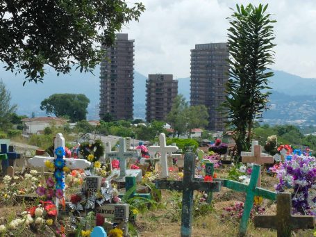 Blick auf ein Friedhof mit bunten Kreuzen und modernen Gebäuden im Hintergrund.