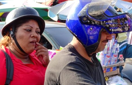 Frau in rotem Shirt und Mann mit blauer Helmkappe auf einem Motorrad in urbaner Umgebung.