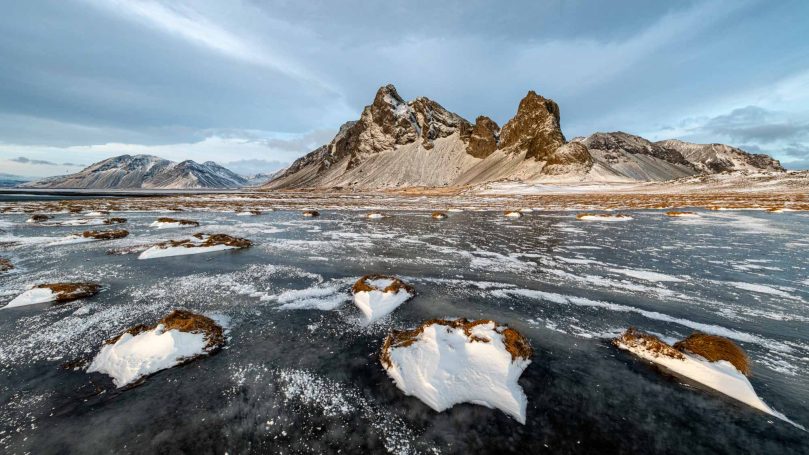 Winterlandschaft mit gefrorenem See, schneebedeckten Bergen im Hintergrund.