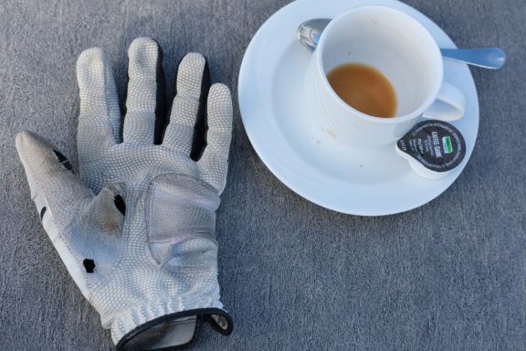 Graue Handschuh neben einer Tasse Kaffee mit einem kleinen Cremebeutel.