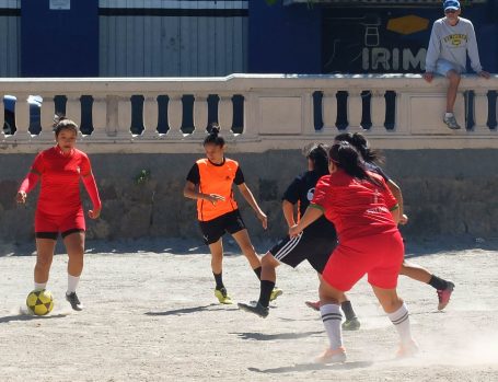 Fußballspiel mit Spielerinnen in orange, schwarz und rot auf einem sandigen Platz.