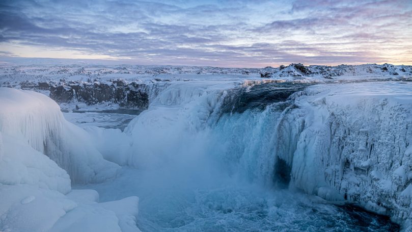 Gefrorener Wasserfall mit eisigem Wasser und stimmungsvollem Himmel bei Sonnenuntergang.